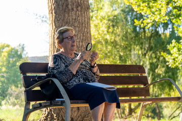 Fototapeta premium enior woman looking at a green leaf with a magnifying glass while sitting on a park bench.
