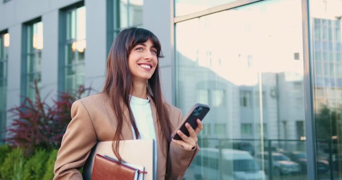 Young Caucasian woman confidently walking near a modern glass office building, smiling as she carries a laptop and a notebook in her hands, while checking her smartphone.