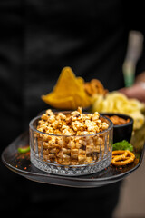 Assorted snacks on a black tray including caramel popcorn, tortilla chips, pretzels, and potato crisps. Close-up, selective focus. Ideal for party, catering, or food photography.