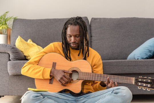 African american man with dreadlocks learning to play guitar sitting on the floor at home - Powered by Adobe