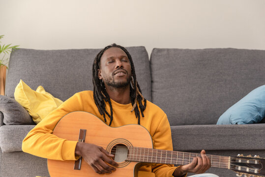 African american man with dreadlocks and closed eyes practicing guitar chords at home