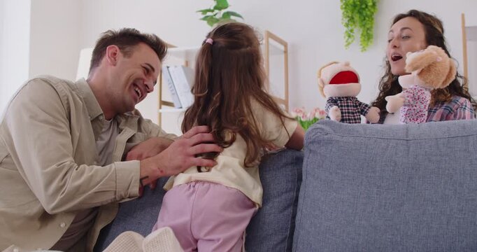 Happy playful family at home play puppet theater show with child girl. Parents and daughter animate plush characters on sofa, laughing together indoors. Family bonding and leisure concept.