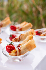 Mini crispy rolls in clear glass bowls with glossy sauce and fresh cherry tomato on white serving tray.