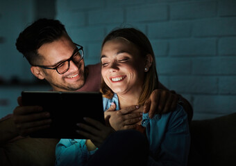 Portrait of young happy couple using tablet computer at night, using modern technology having fun with glowing screen in dark office or at home, watching tv together at home. Shot of a couple resting