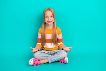 Cheerful little girl sitting cross-legged on teal background wearing trendy striped sweater and pink sneakers
