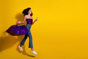 Young woman jumps with purple shopping bags smiles on her phone against a bright yellow background