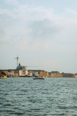 Tranquil cityscape. Historic Venetian buildings line canal beneath soft sky, with a construction crane visible above. Lone boat rests on still water. Architectural detail highlighted by gentle light.