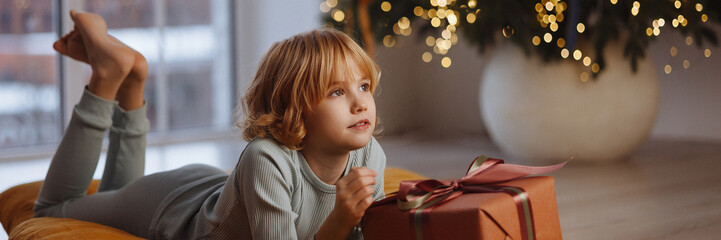 Happy Caucasian girl, 9 years old, blond curly hair, in pajamas, lying on soft cushion holding gift box near Christmas tree with lights and white wall. Concept of holiday, New Year, childhood joy.