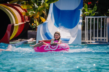 A smiling young girl rides a pink inflatable tube into the pool after sliding down a blue and white...
