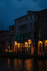 Captivating view of historic Venetian architecture along a canal, bathed in the warm glow of evening lights. Ornate facade reflects beautifully on the water, creating an enchanting and timeless scene.