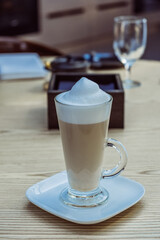 Glass of creamy latte topped with frothy milk foam sits elegantly on a white plate, surrounded by a cozy cafe atmosphere with wooden table and soft lighting