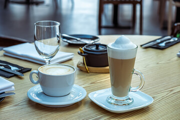 Two coffee beverages on a wooden table, featuring a latte in a white cup and a frothy cappuccino in a glass, surrounded by elegant dining utensils and a cozy atmosphere