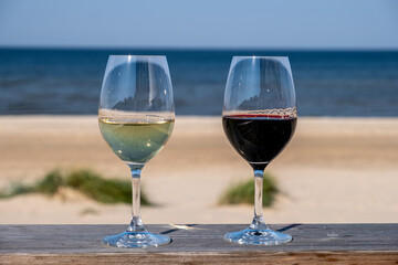 Two elegant wine glasses, one filled with white wine and the other with red wine, placed on a wooden table overlooking a serene beach and calm ocean waves