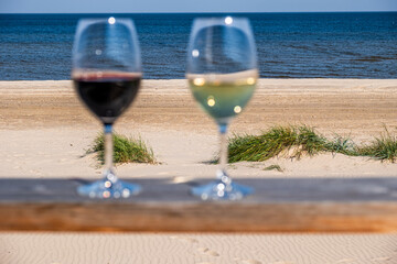 Two elegant wine glasses filled with red and white wine are placed on a wooden table overlooking a serene beach, capturing a moment of relaxation and enjoyment by the sea