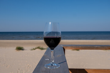 Glass of red wine placed on wooden railing overlooking serene beach with calm ocean waves and clear blue sky, creating a relaxing atmosphere for leisure moments