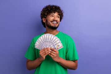 Smiling young man holding cash fan in his hands against a vibrant purple background expressing joy...