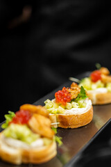 Gourmet appetizers on toasted baguette with creamy spread, grilled meat, red caviar, herbs, and vegetables. Elegant fine dining presentation on dark tray.