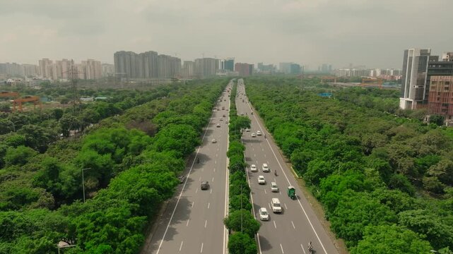Aerial view of the Noida-Greater Noida Expressway shows a ribbon of grey asphalt cutting through lush green trees, with the skyline of buildings in the distance, Noida, Uttar Pradesh, India.