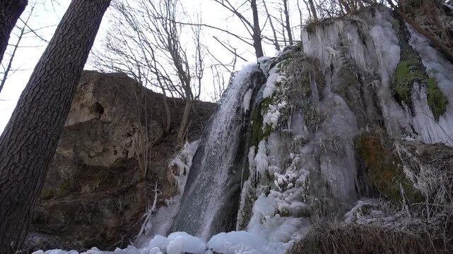 A frozen waterfall on a mountain river. The Bigar waterfall below Stara Planina is bound by ice, you can see ice stalactites and other ice forms, and some unfrozen water falling. The camera moves down