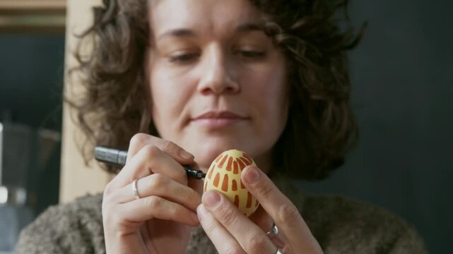 Close up of woman with short curly hair holding black marker and drawing on yellow colored egg with orange pattern