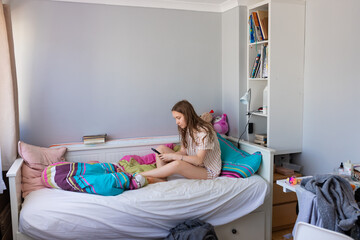 A teenage girl in pajamas lies in bed with her mobile phone in a messy room