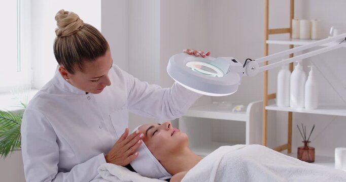 Female professional cosmetologist doing skin care facial treatment examining its condition for woman client with special lamp in beauty clinic. Cosmetology and beauty procedure concept. 4k video.