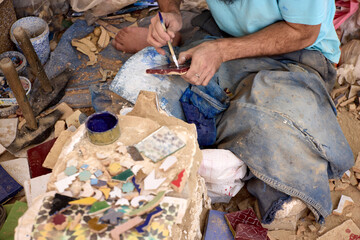 A skilled artisan carefully paints a mosaic tile in a ceramics school located in Fez, Morocco. Surrounding him are colorful pieces of ceramic and tools, highlighting traditional craftsmanship