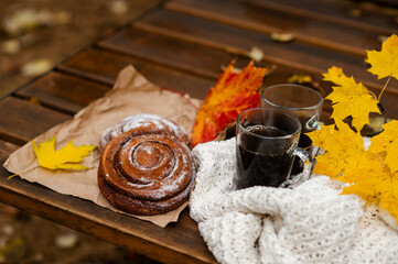 Cozy autumn picnic scene with two cups of coffee, cinnamon rolls and colorful maple leaves on a wooden bench wrapped in a white knitted blanket