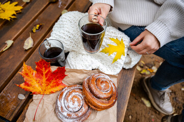 Partial view of woman in white knitted sweater holding cup of coffee at wooden bench with pastries and autumn leaves
