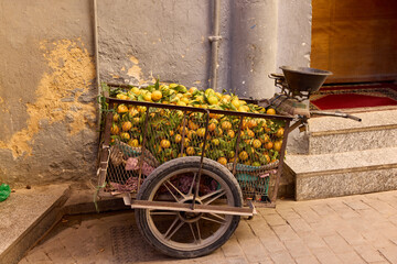 A cart overflowing with fresh fruits stands on a cobblestone street in Morocco. The warm midday sun highlights the vibrant colors of the produce, capturing the essence of local market culture