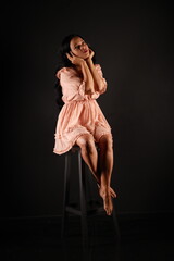 Long-legged barefoot dark-haired woman in a beige dress sitting on a chair on a black background