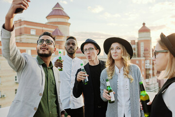 Young Middle Eastern man taking selfie on smartphone with his trendy male and female friends hanging out on rooftop terrace with beer