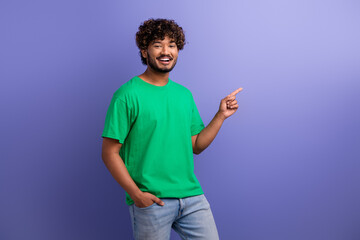 Cheerful young man gesturing to the side in bright green t-shirt and jeans against a vibrant purple...