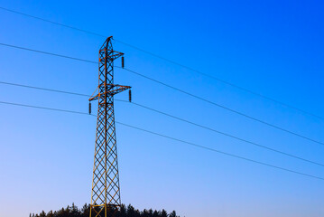 Tall metal power line tower stands against a clear blue sky near trees in a rural area during evening light