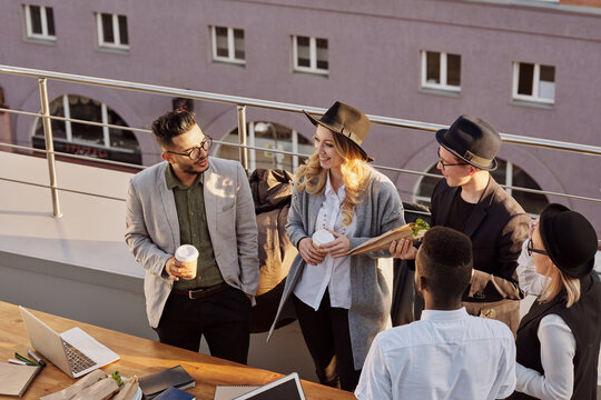 Multiethnic group of stylish young male and female coworkers chatting, drinking coffee and eating snacks on rooftop terrace, high angle view - Powered by Adobe