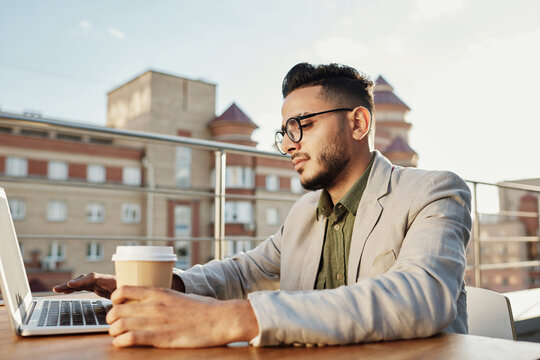 Young handsome Middle Eastern man with glasses sitting at table on rooftop terrace and working on laptop