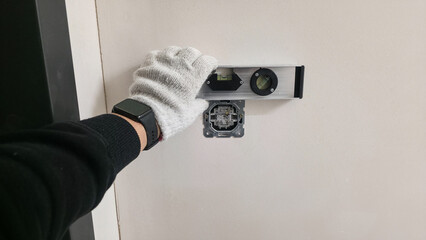 Electrician aligning wall socket using spirit level. Gloved hand holds tool for precise positioning before installation. Professional electrical work in progress.