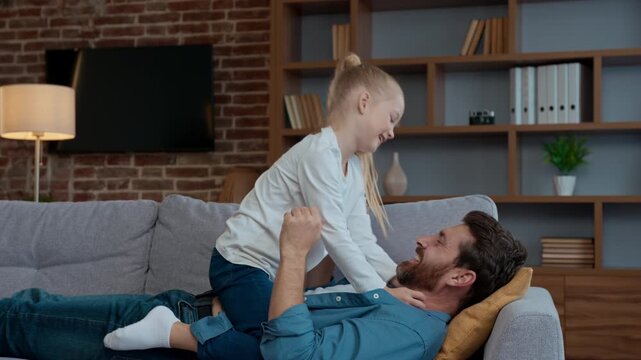 Caucasian happy family at home playing tickling father with daughter laughing tickle man dad lying on sofa couch play with loving child kid little girl fooling having fun enjoying weekend together