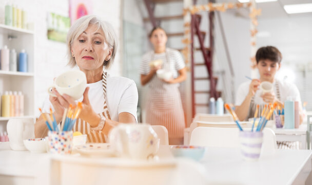 Experienced interested mature female artist sitting at table in pottery workshop, painting designs on ceramic cup. Art craft activity concept..