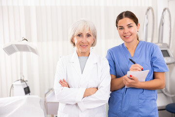 Senior woman cosmetologist and her assistant nurse smiling and looking at camera in clinic of...