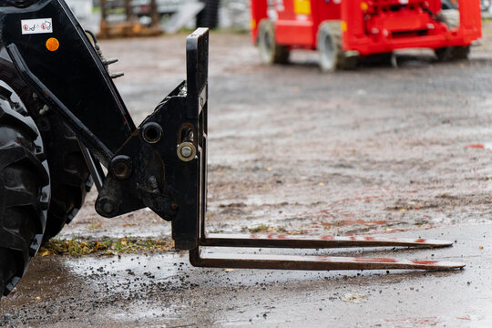 Close view of forklift attachment mounted on front loader tractor with metal pallet forks on wet construction site ground representing industrial lifting, logistics and heavy equipment operation