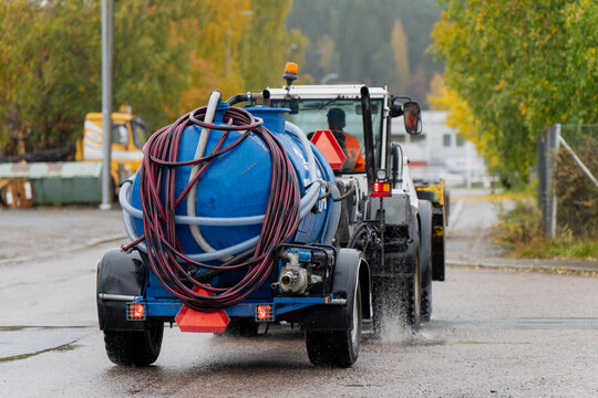 Utility tractor towing blue vacuum sanitation tank with hoses and spray system driving on wet road surface, used for wastewater suction, septic maintenance and urban environmental cleaning tasks