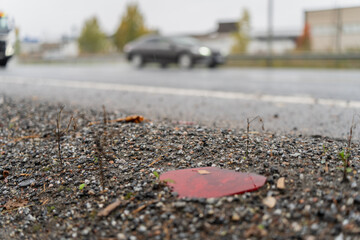 Broken red road reflector lying on gravel shoulder near wet asphalt highway with moving car in background, illustrating traffic infrastructure detail, vehicle safety, and maintenance condition of road