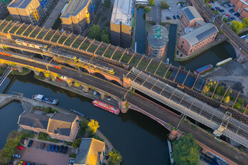 Top-down sunrise aerial of Manchester’s Castlefield viaducts showing trams, a train, and canal boats along Bridgewater Canal
