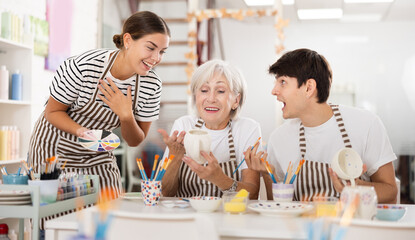 Enthusiastic senior female teacher sharing experience pottery craftsmanship, assisting happy young guy and girl, painting ceramic mugs and plates at table in workshop