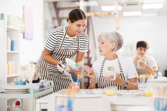 Pleasant young female instructor sharing art of ceramic dishware painting with interested enthusiastic mature woman participating pottery master class - Powered by Adobe