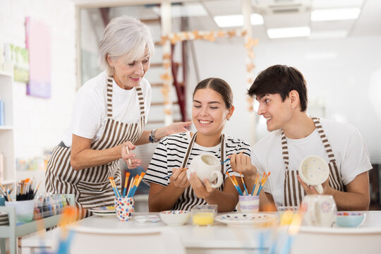 Friendly older mother and young adult daughter and son, wearing aprons, talking cheerfully while working on painting ceramic dishware in family pottery workshop. Generational collaboration concept..
