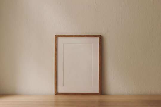 Blank vertical wooden picture frame poster mockup on wooden table desk leaning against beige wall. Artistic display template. Minimalist interior. Natural light. Neutral design. Empty photo frame.