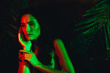 Portrait of a young woman in vibrant neon lights surrounded by tropical leaves, highlighting beauty...