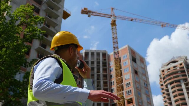 Close up of handsome male worker in hard hat talking in walkie-talkie standing at construction site on street outdoor. Low angle view of man constructor engineer using receiver at work
